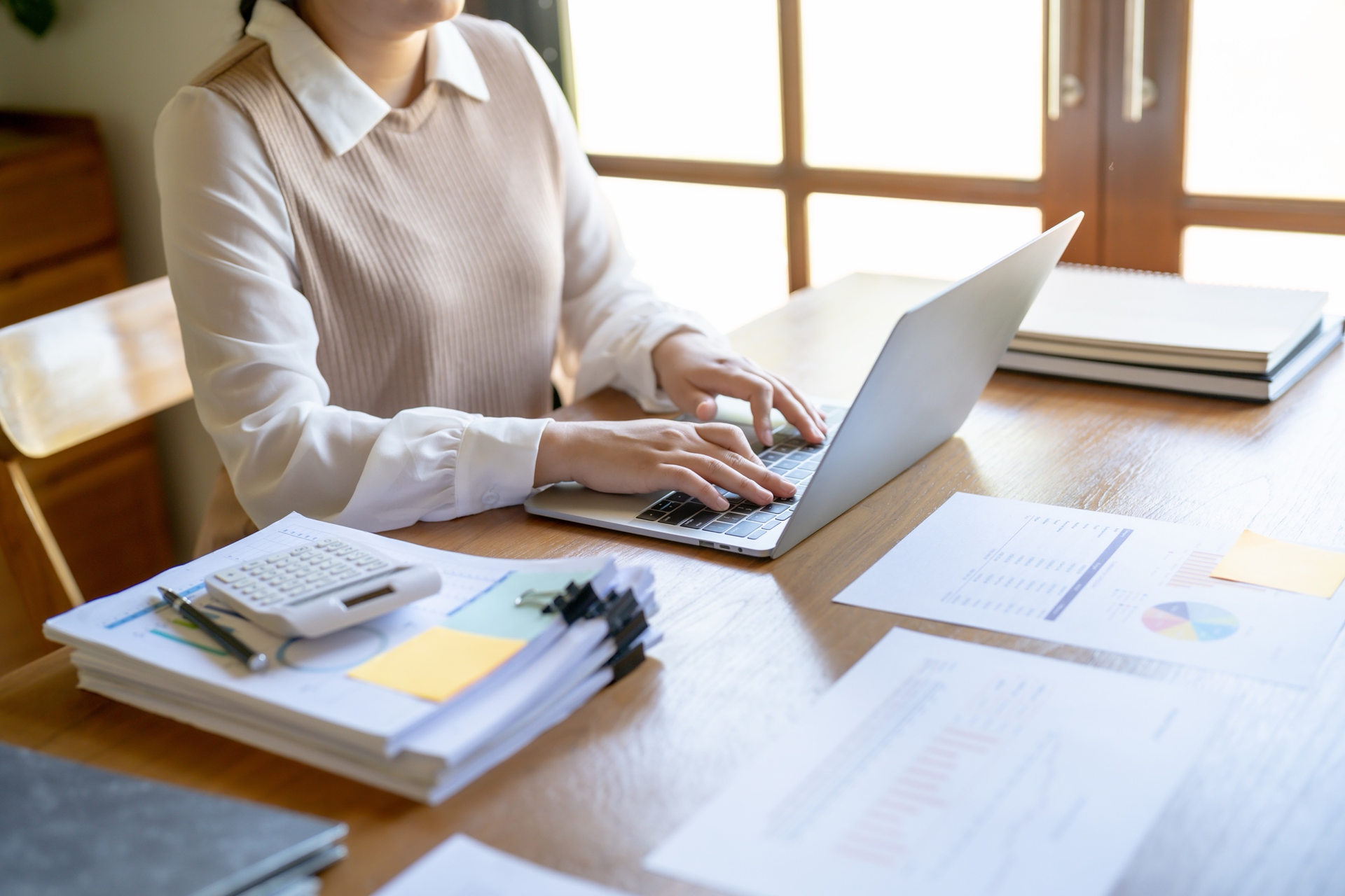 Businesswoman doing small business accounting at a laptop with stacks of paperwork